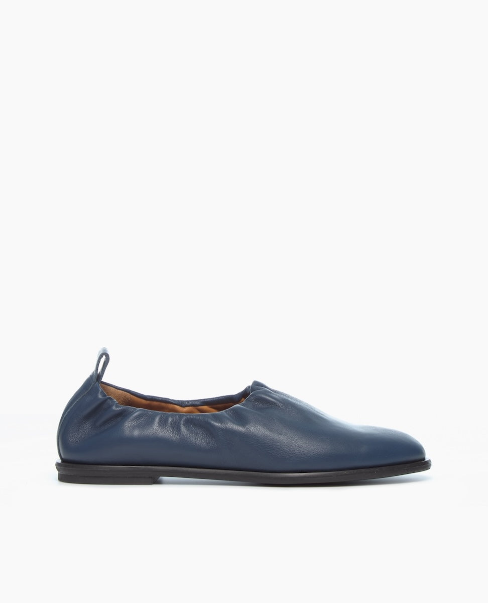 A single blue leather slip-on shoe with a minimalist design and a flat black sole is displayed against a white background.