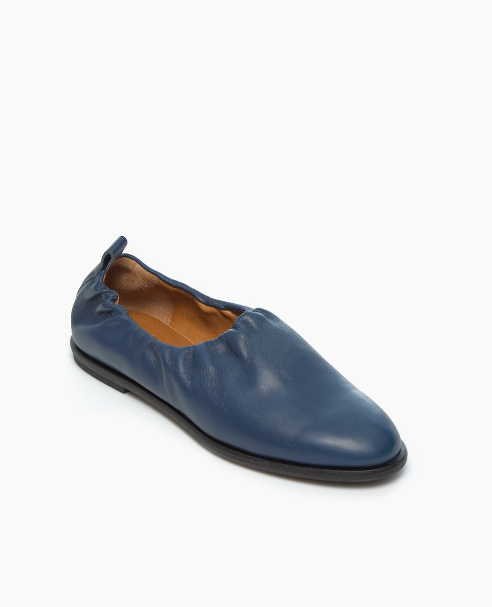 A single blue leather flat shoe with a slightly gathered design around the opening and a black sole is displayed against a plain white background.
