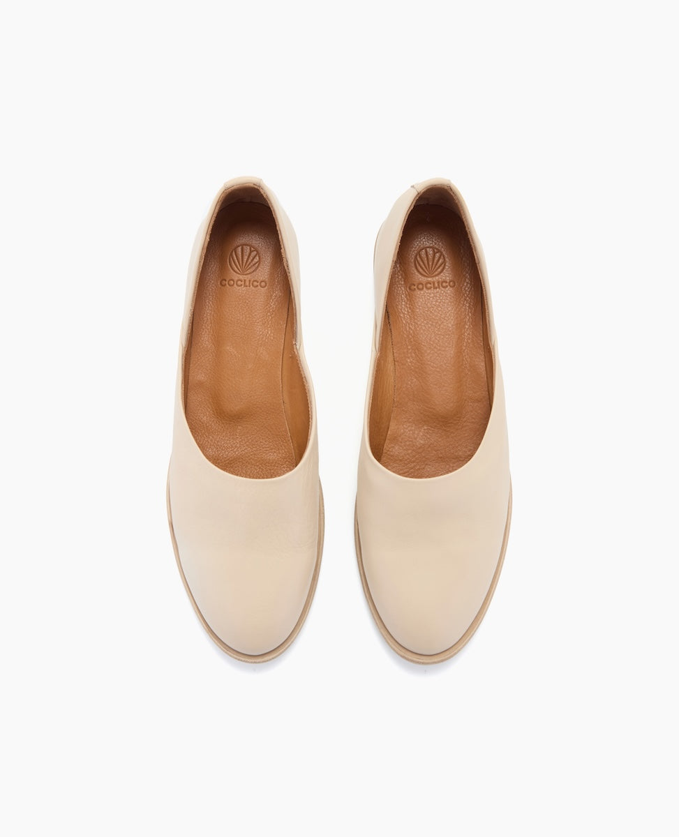 A pair of beige slip-on shoes with a smooth, rounded toe, featuring a tan leather interior and a subtle logo on the insole.