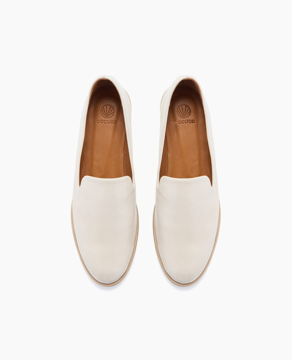 A pair of sleek, white slip-on shoes with a smooth leather texture, featuring a rounded toe, tan interior lining, and a subtle logo on the insole.
