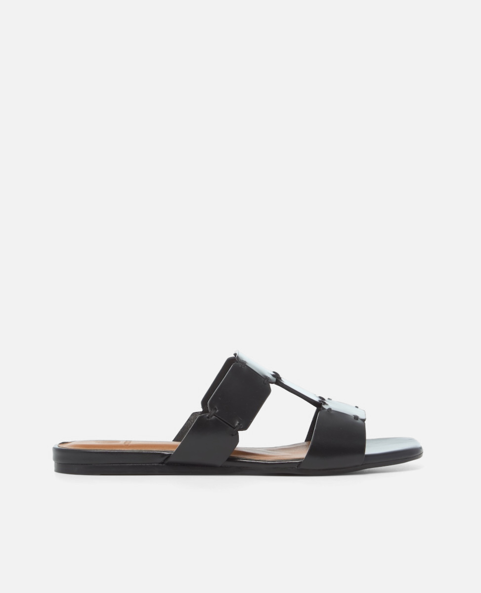 Side view of a black vachetta leather T-strap slide sandal with octagonal hand stitched panels, square toe, and flat sole, on a white background.