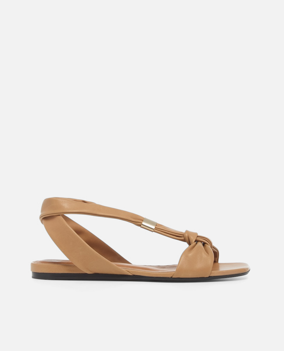 Side view of a tan leather flat sandal with tubular straps, an adjustable brushed-metal keeper at the center strap, toe knot detail, and a square toe, on a white background.