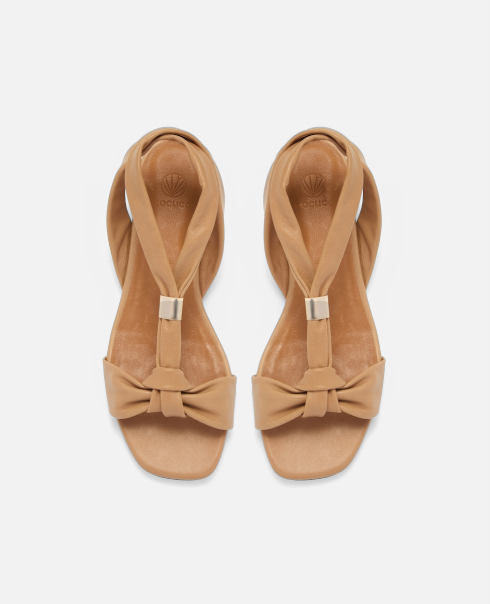 Top view of a tan leather flat sandal with tubular straps, an adjustable brushed-metal keeper at the center strap, toe knot detail, and a square toe, on a white background.