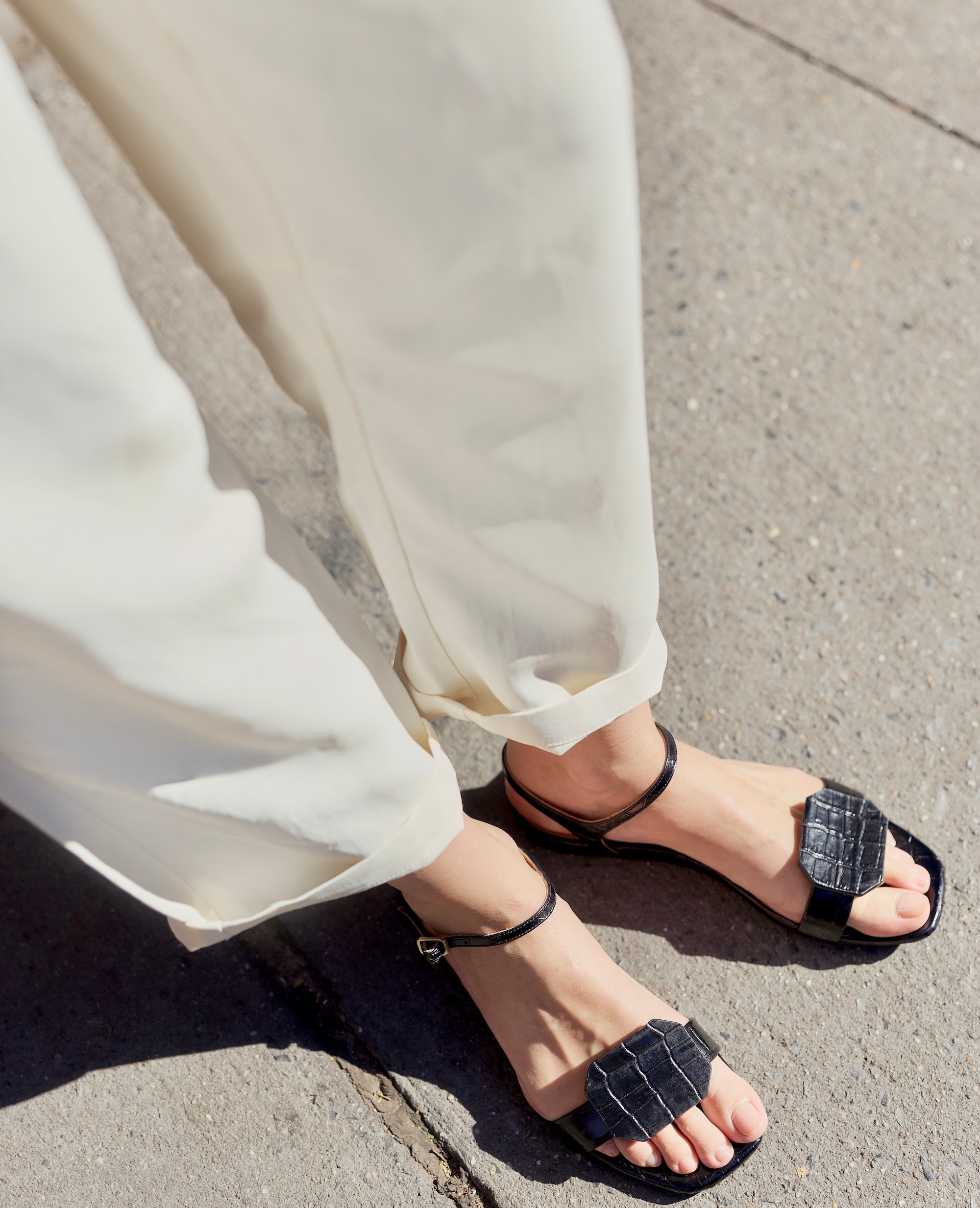 Person wearing black croco sandals with a croc textured strap on a city sidewalk
