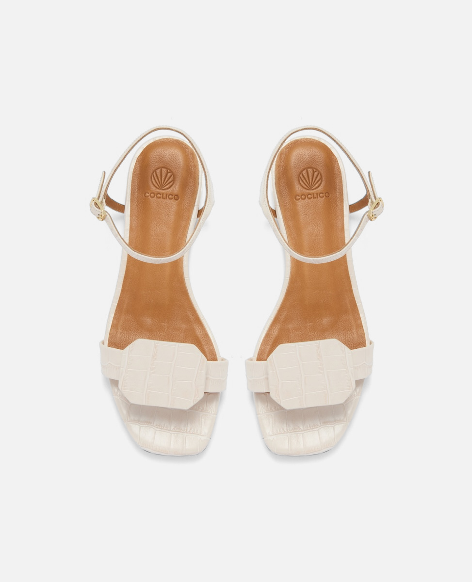 Top view of a bone-colored croc-embossed leather flat sandal with a slim ankle strap and a octagonal accent on the front strap, square toe, on a white background.