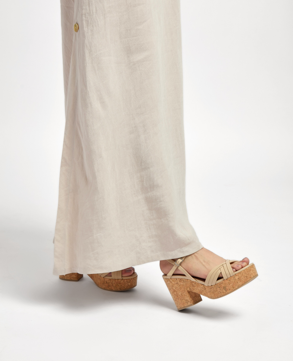 Beige platform sandals with cork wedge heels, featuring crisscrossing straps and an open-toe design, paired with a long, light-colored linen skirt.