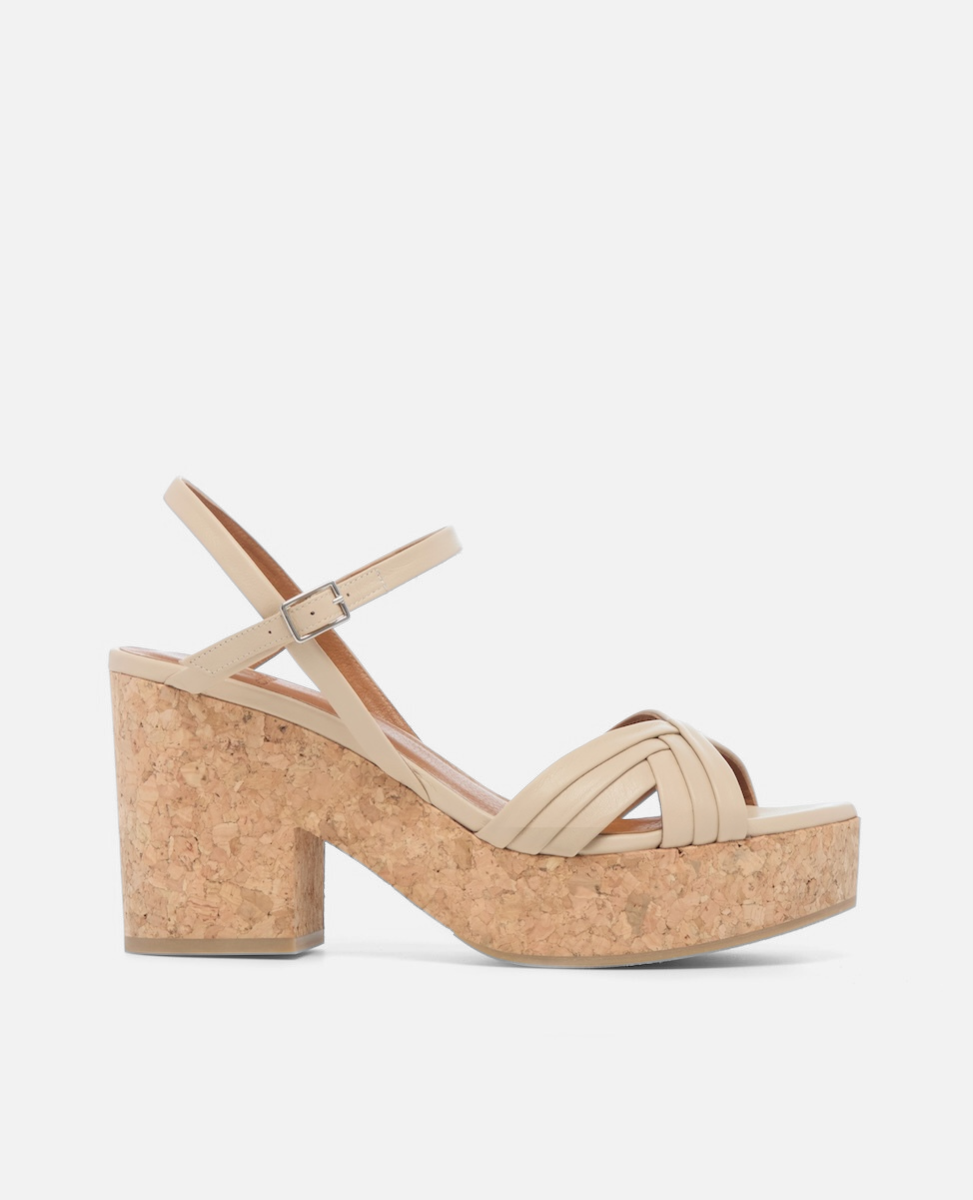 A beige sandal with a cork platform heel features crisscrossing straps over the toe, an adjustable ankle strap with a silver buckle, and a chunky, cork heel.