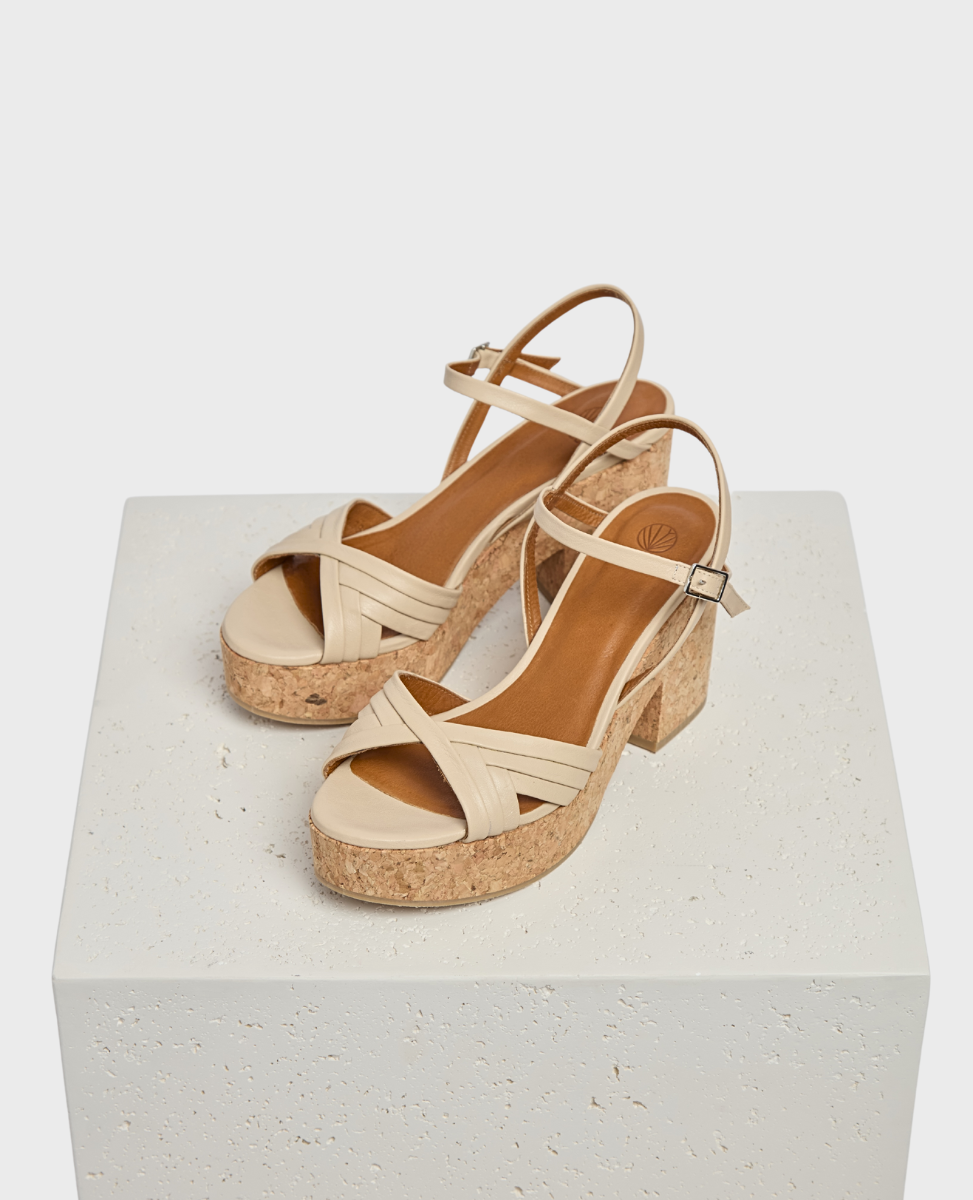 Beige strappy sandals with a cork block heel, featuring a crisscross design on the front and an adjustable ankle strap, resting on a textured white platform.