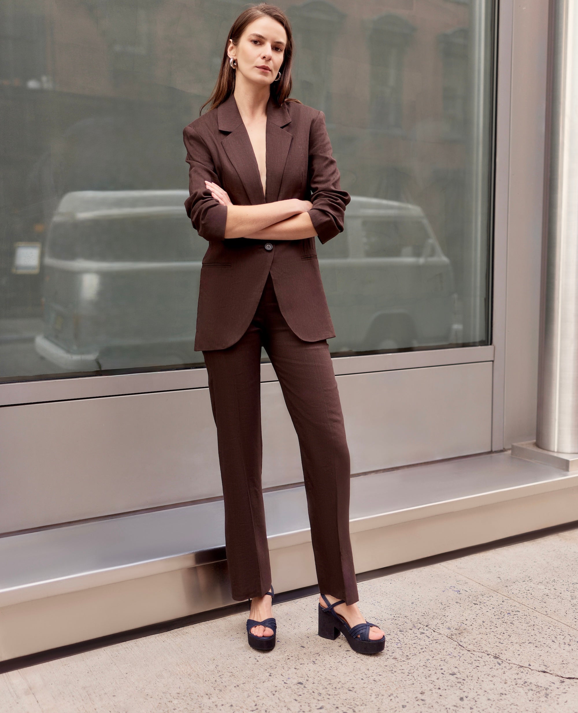 Woman in a brown suit standing in front of a glass window with a city street reflection, wearing a brown suit and platform cork navy sandals