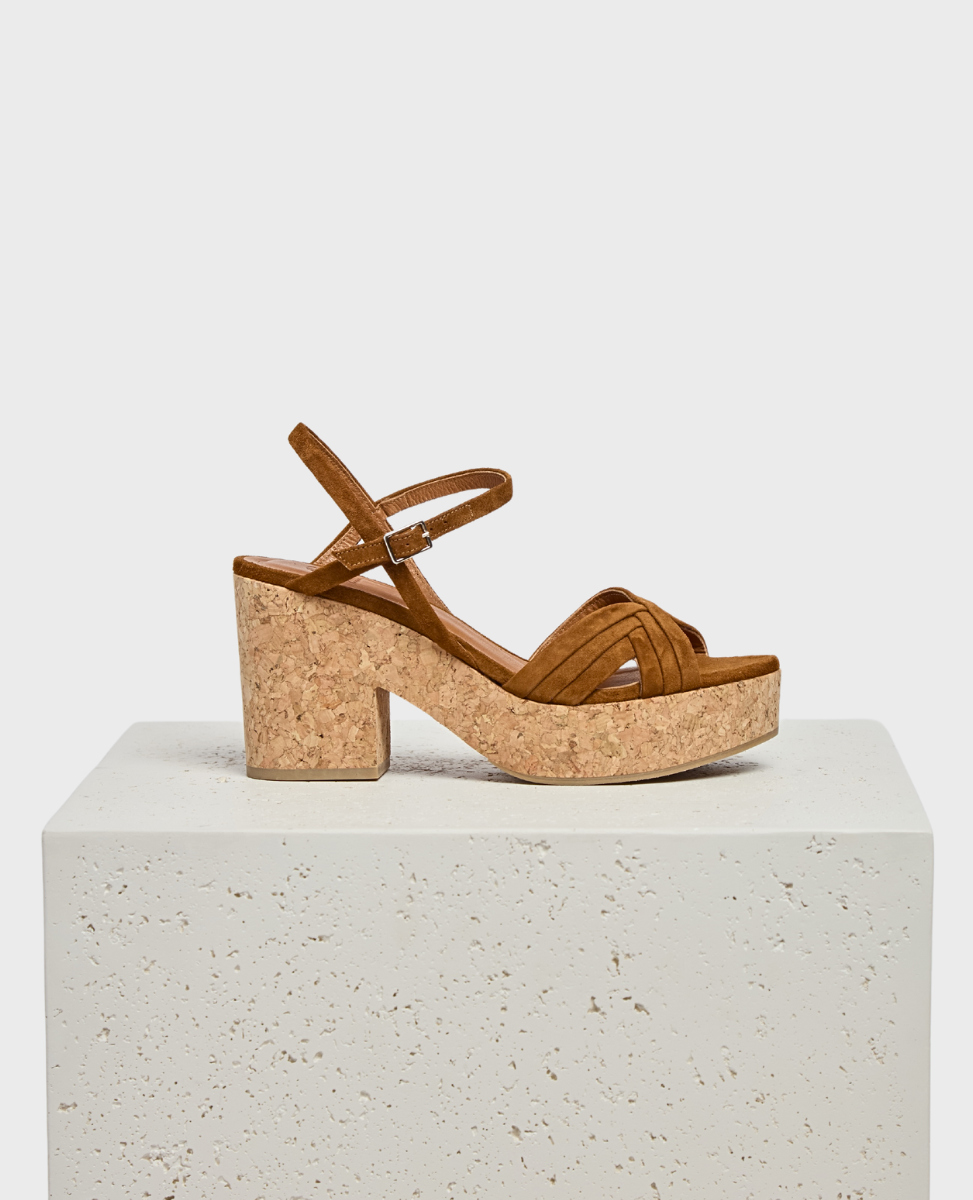 A brown sandal with a cork block heel and platform, featuring a strappy suede upper and an adjustable ankle strap, displayed on a textured white pedestal.