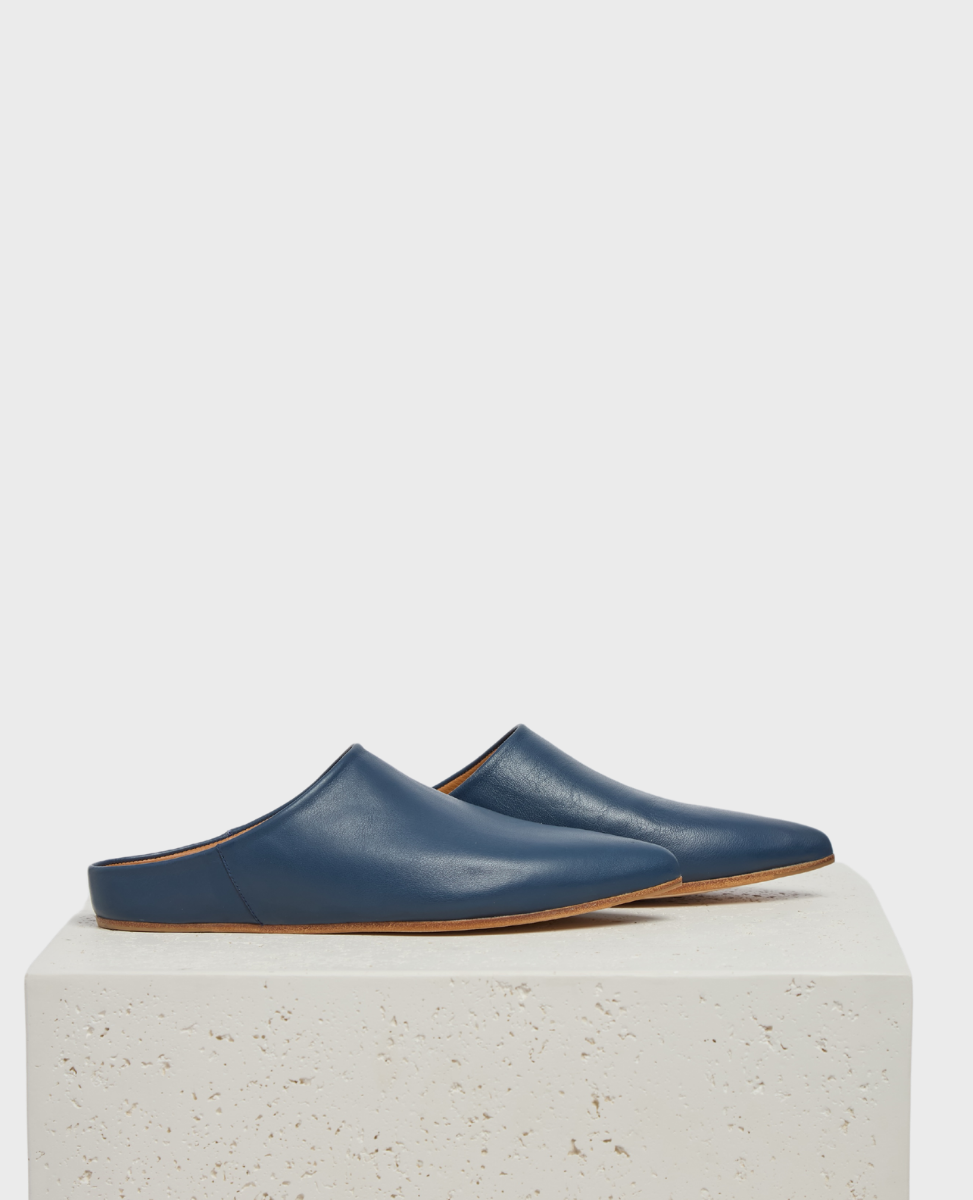 A pair of sleek, blue leather slip-on mules with a pointed toe and flat brown sole, displayed on a textured, light-colored stone surface.