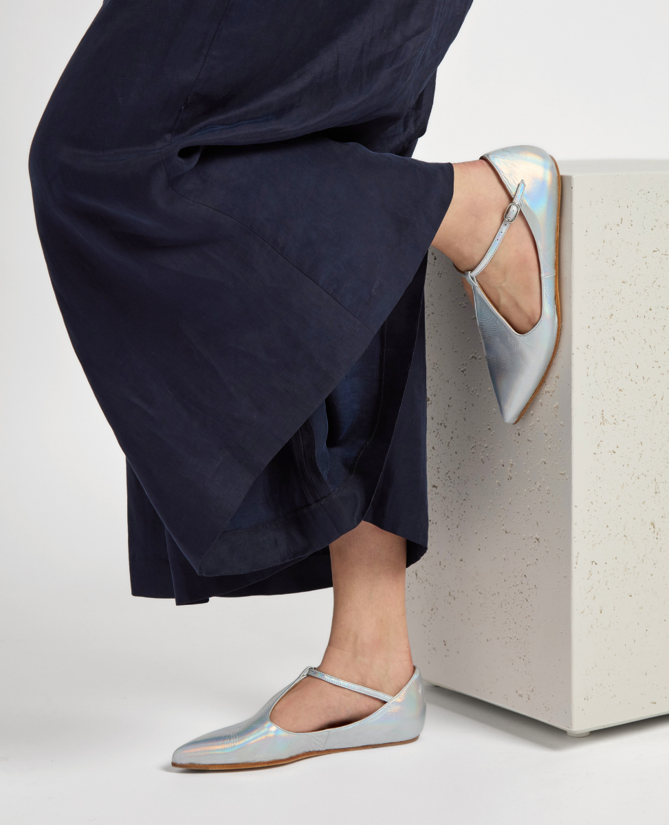 A pair of sleek, metallic silver ballet flats with a pointed toe, thin ankle strap, and a smooth, shiny texture, worn by a person in dark, flowing pants.