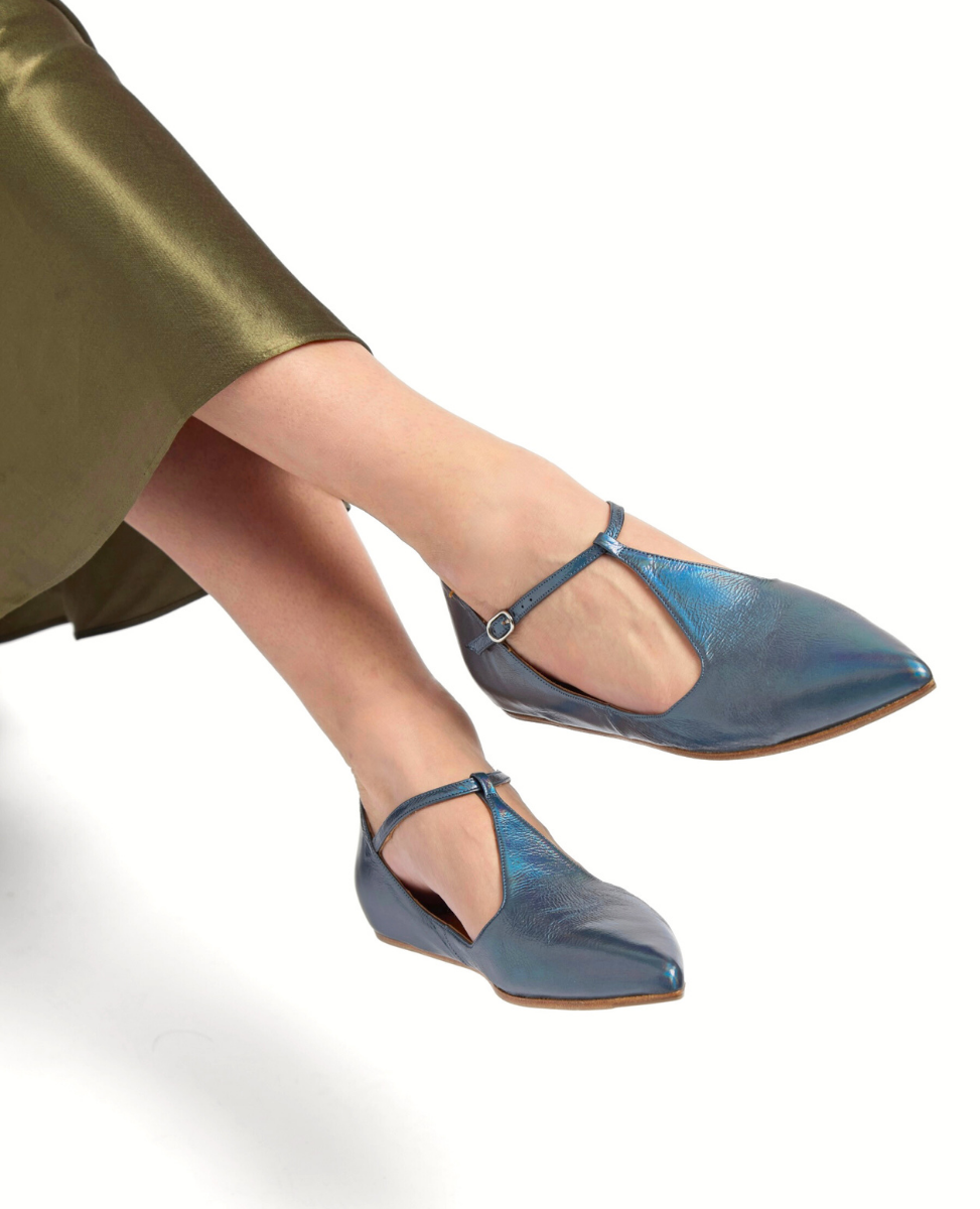 A pair of sleek, metallic blue T-strap flats with a pointed toe, smooth leather texture, and a small buckle on the strap, worn with a green satin skirt.