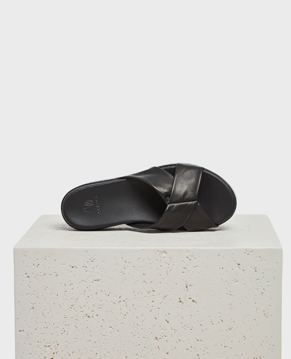 A sleek black slide sandal with a smooth leather crisscross strap design, featuring a flat sole and subtle embossed logo on the footbed, resting on a textured stone block.