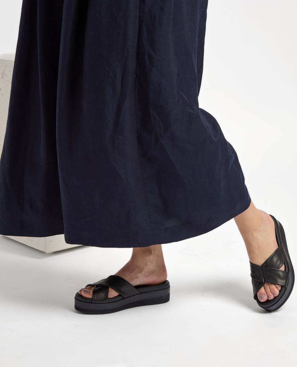 A pair of black leather sandals with wide crisscross straps and a thick, flat sole, worn by a person in a long, dark blue skirt.