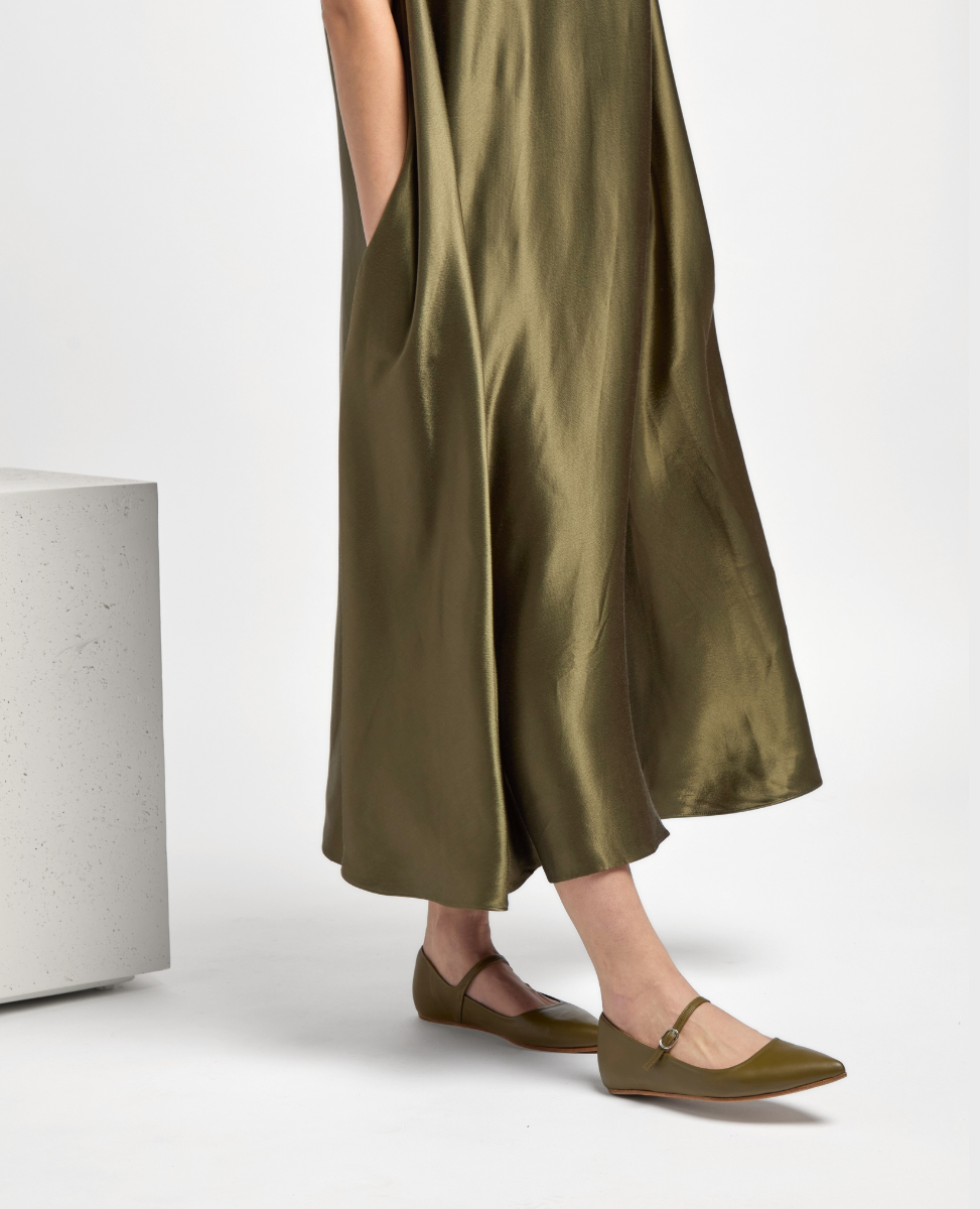 A pair of sleek, olive green, pointed-toe flats with a smooth, shiny texture and a thin ankle strap, worn with a flowing, matching satin dress.