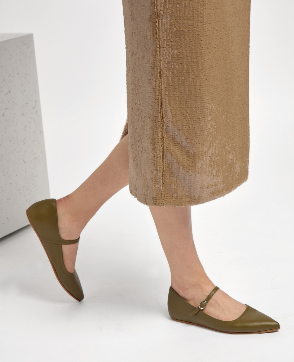 Olive green, pointed-toe flats with a smooth leather finish, featuring a thin strap with a small buckle across the top, paired with a textured, light brown skirt.