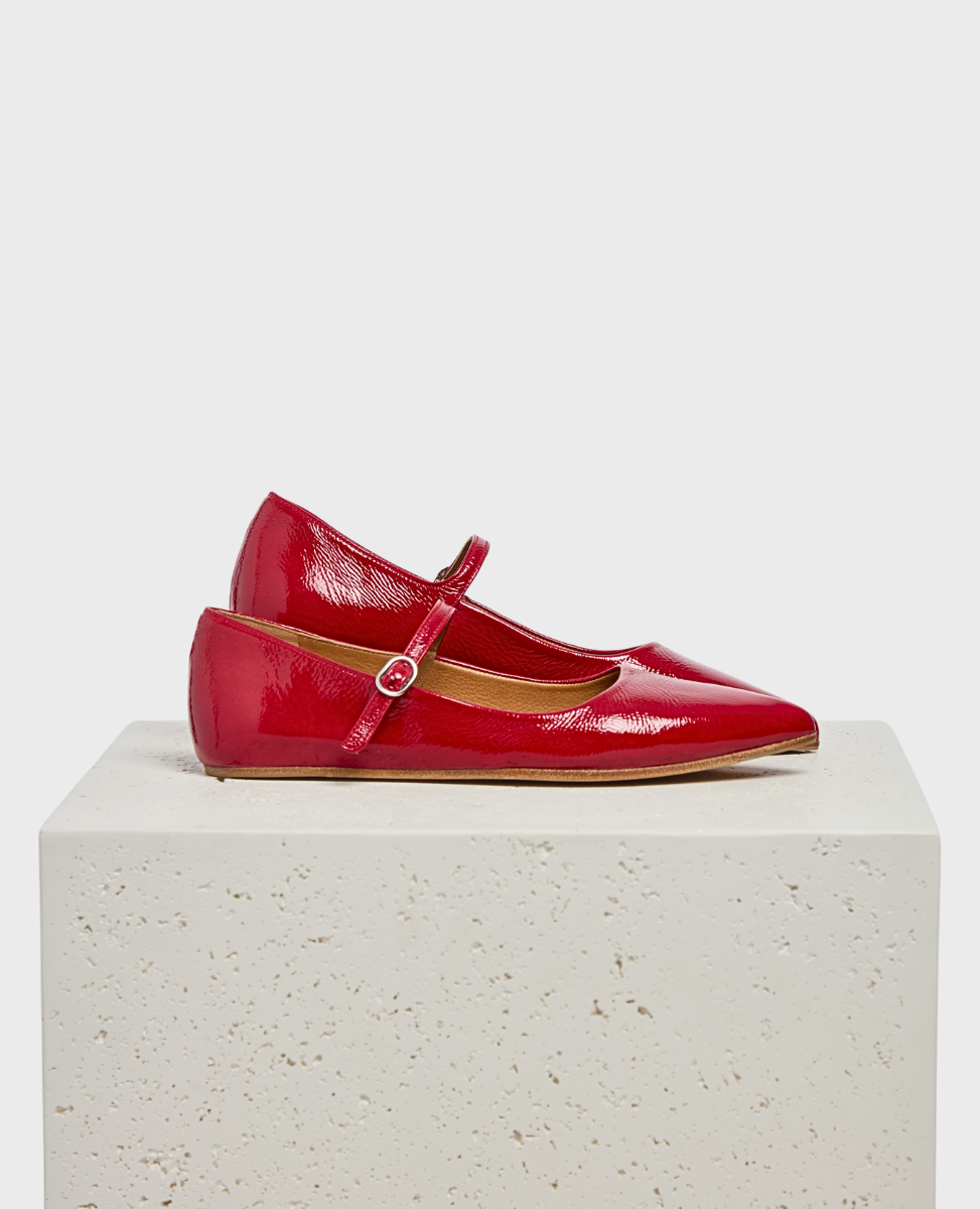 A pair of glossy red patent leather Mary Jane flats with pointed toes, a single strap with a small buckle, and a flat sole, displayed on a textured white pedestal.