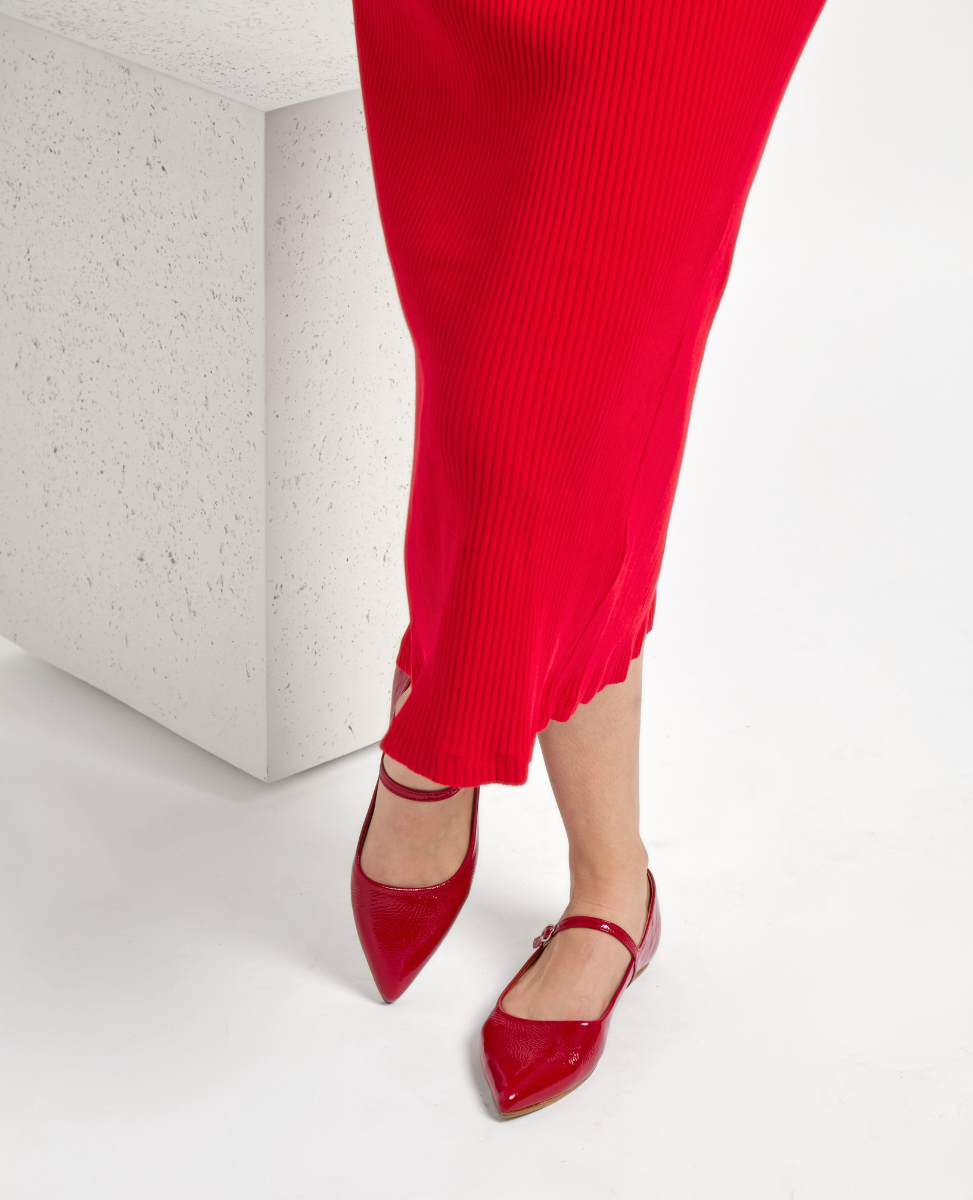 A pair of shiny red patent leather pointed-toe flats with a low heel and a single strap across the top, worn with a long, ribbed red skirt.