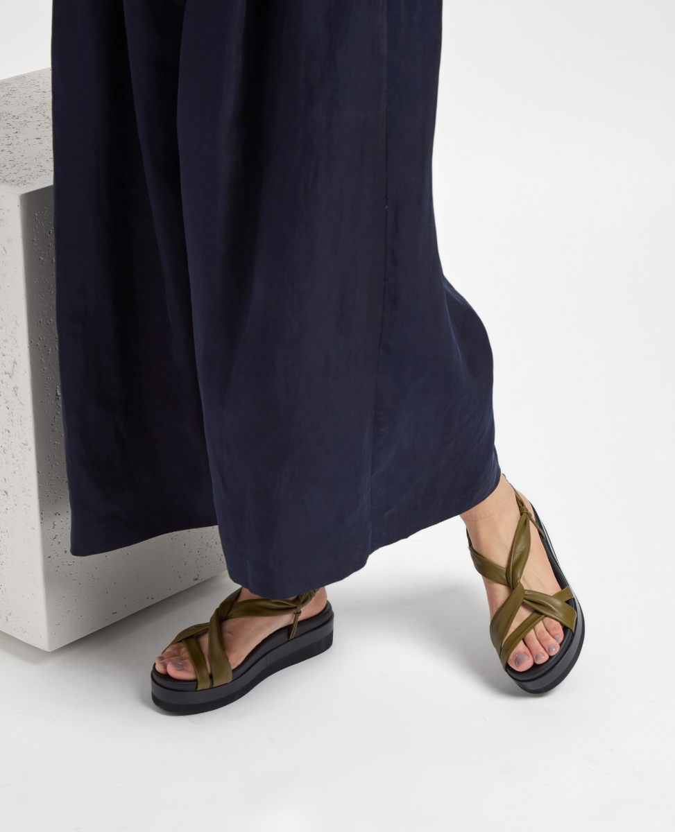 A pair of sandals with olive green, crisscrossing leather straps and a thick black sole, worn by a person in dark blue wide-legged pants.