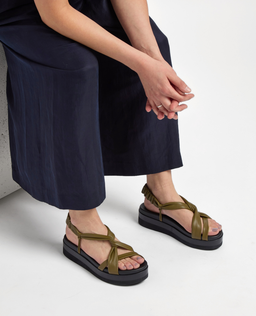 The product is a pair of olive green sandals featuring wide, crisscrossing leather straps and a thick black platform sole, with a smooth texture and an open-toe design.