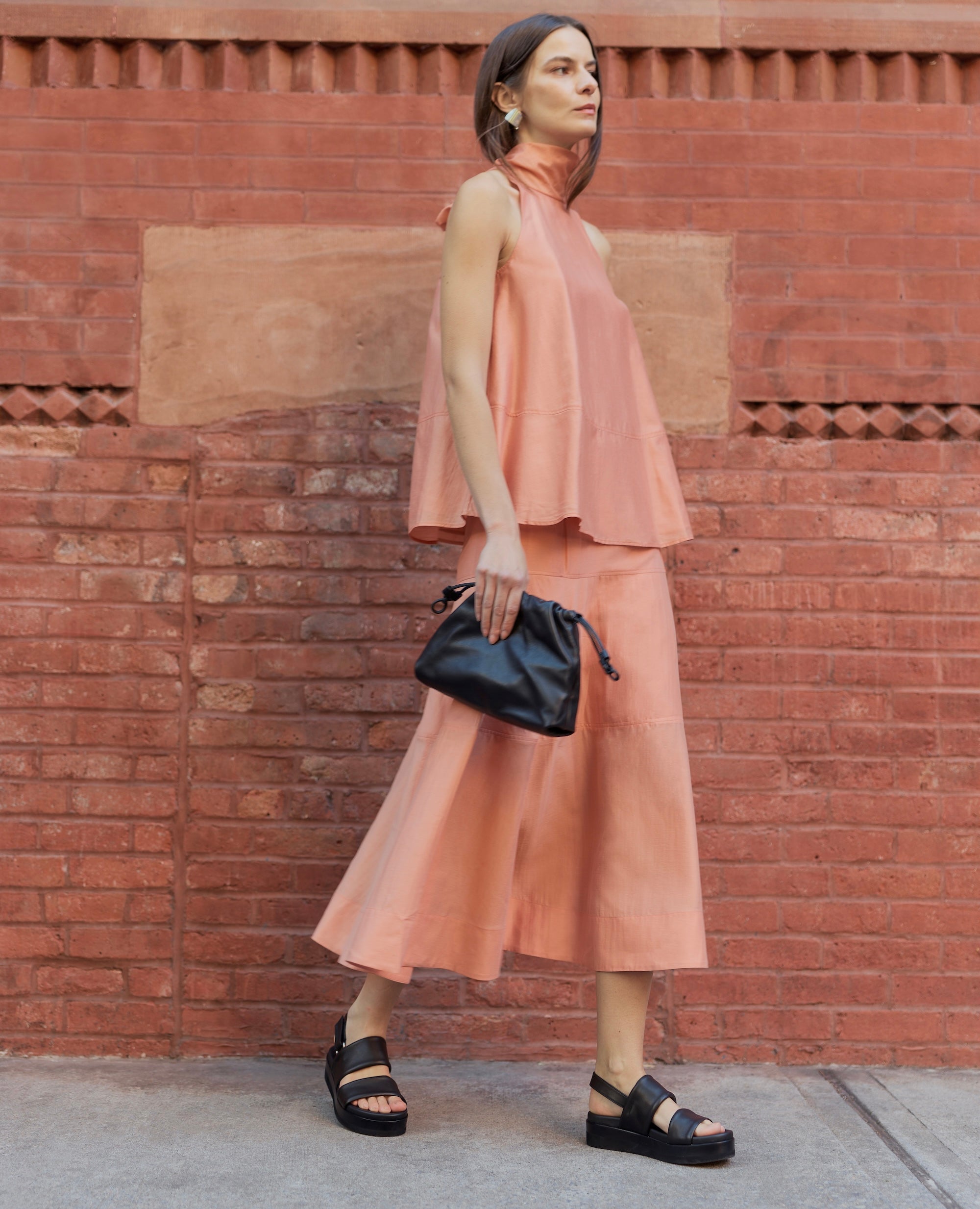 Woman in a peach outfit, black bag, and black tubular leather flatform sandals standing against a brick wall