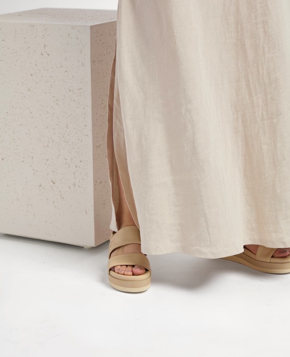 A pair of beige, open-toed sandals with wide straps and a flat sole, worn by a person standing next to a textured, light-colored cube.