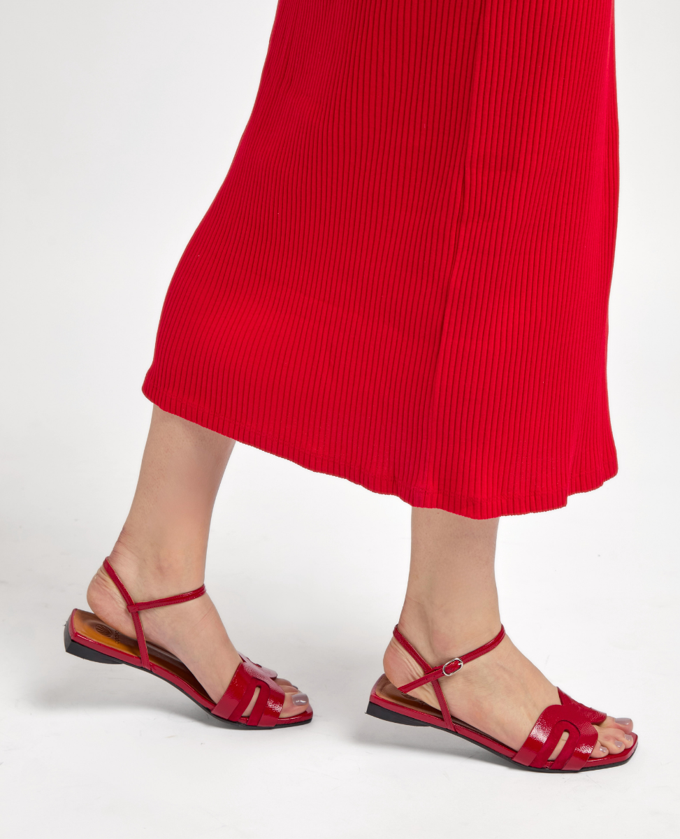 Red sandals with a flat sole, featuring thin straps and a buckle closure, complement a red pleated skirt.