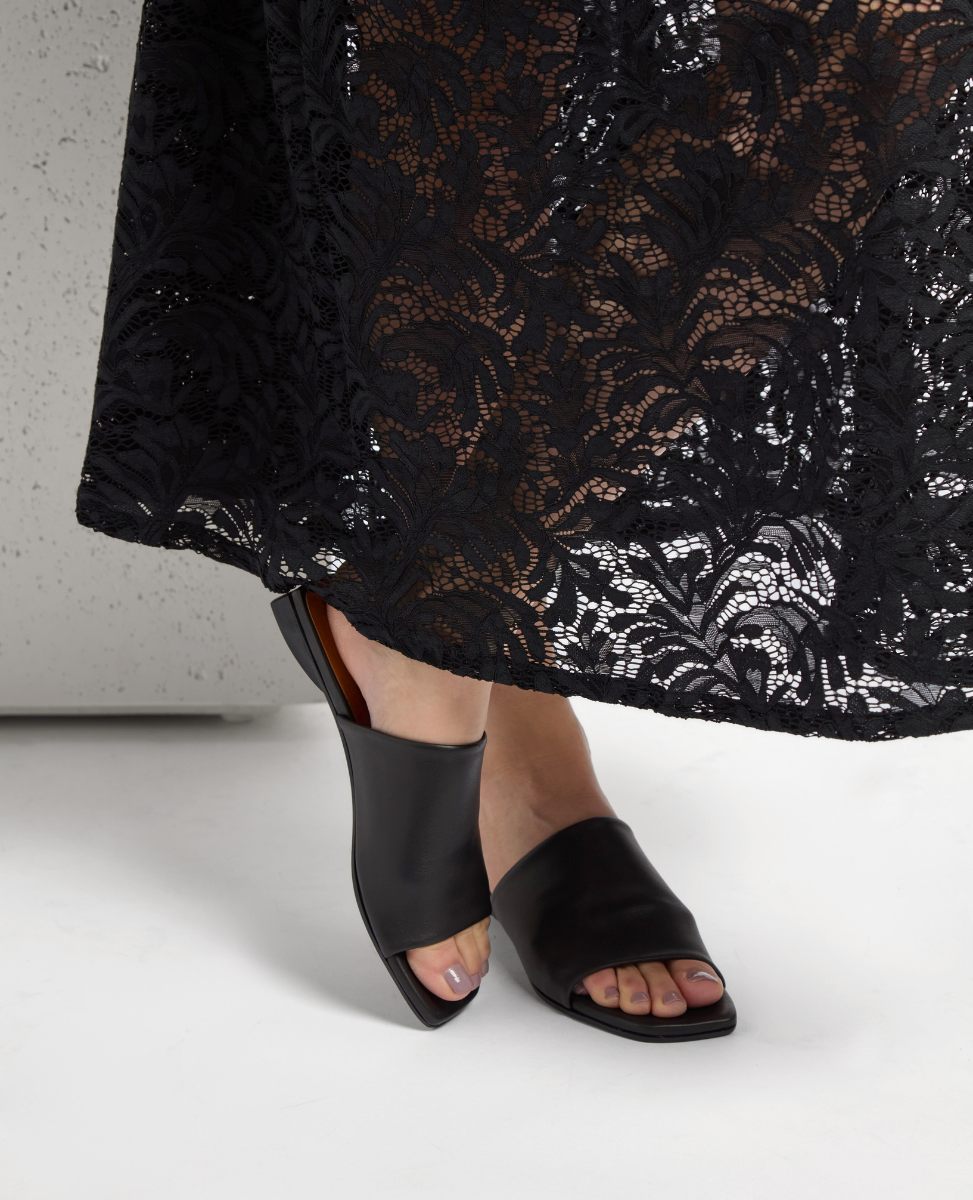 A pair of black, open-toed, slip-on sandals with a sleek leather finish and a low, angular heel, worn with a long, black lace skirt featuring intricate floral patterns.