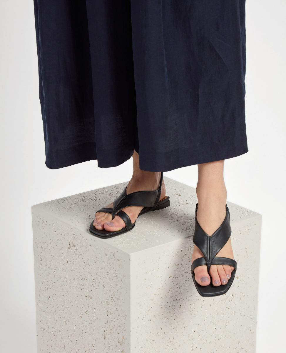 Black leather sandals with a V-shaped strap design, open toe, and flat sole, worn by a person standing on a textured white cube.