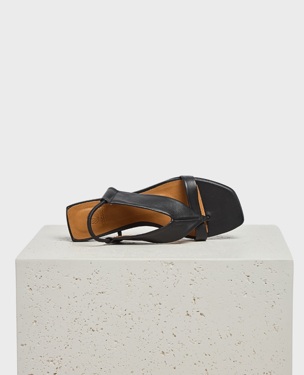 A black leather sandal with a square toe, featuring a minimalist design and a thin ankle strap, displayed on a textured white block.