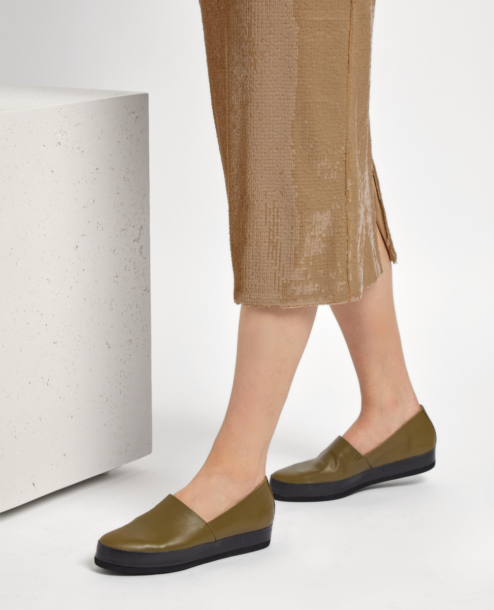 Olive green slip-on shoes with a smooth leather finish, featuring a rounded toe and a contrasting black flat sole, worn by a person in a textured, light brown skirt.