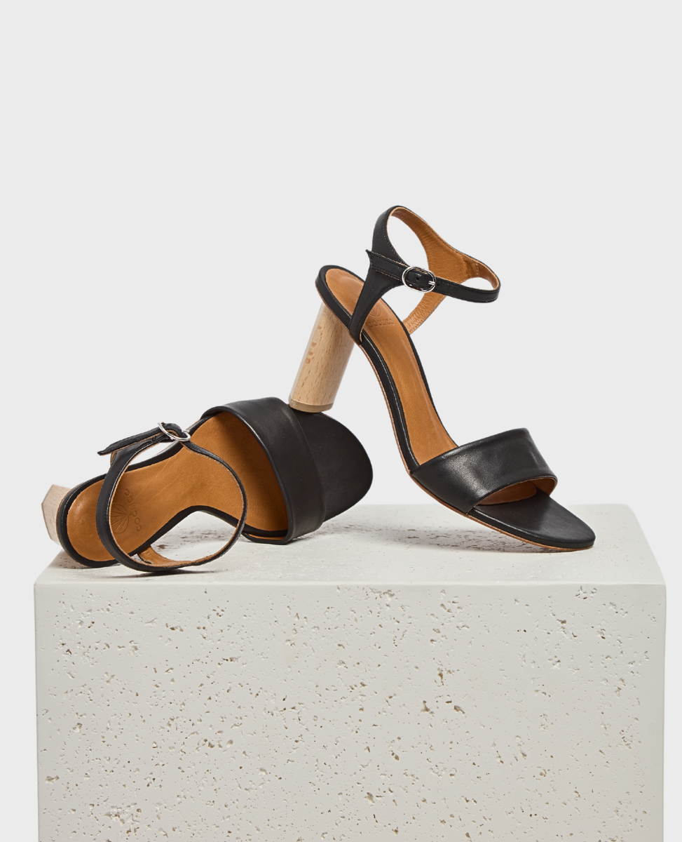 A pair of stylish black leather sandals featuring a unique cylindrical wooden heel, an open toe design, and an adjustable ankle strap with a buckle, resting on a textured white stone surface.