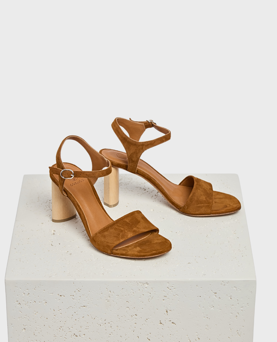 A pair of brown suede sandals with a block heel, featuring an open toe design, ankle strap with a buckle, and a smooth leather insole, resting on a textured white platform.