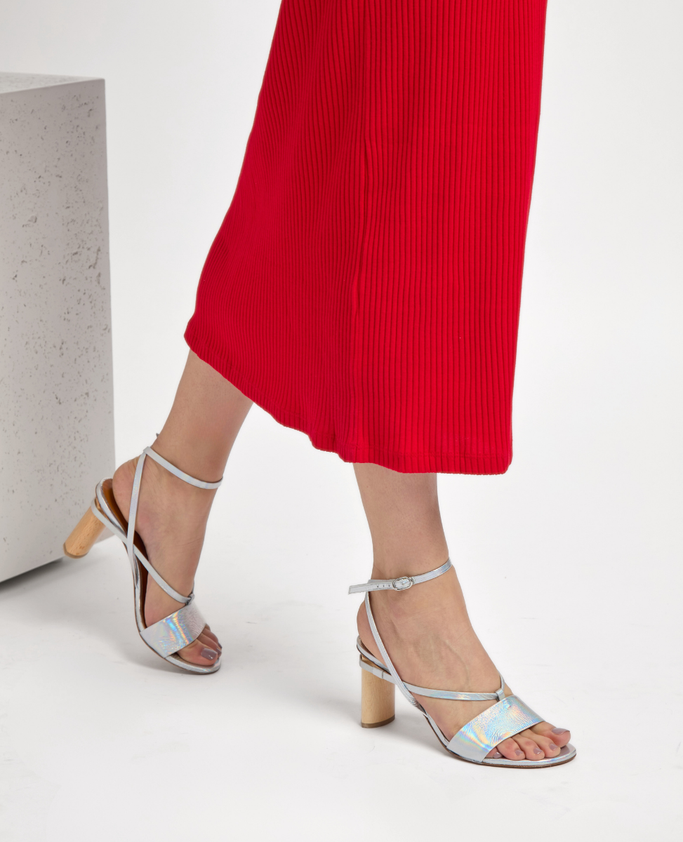 Silver strappy sandals with a holographic sheen, featuring a chunky wooden heel and a delicate ankle strap, paired with a vibrant red pleated skirt.