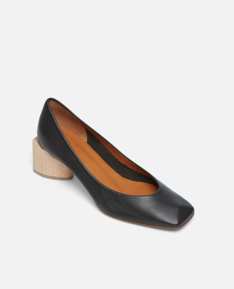 Angled view of a black leather pump with a square toe and a solid architectural, natural wood block heel on a white background