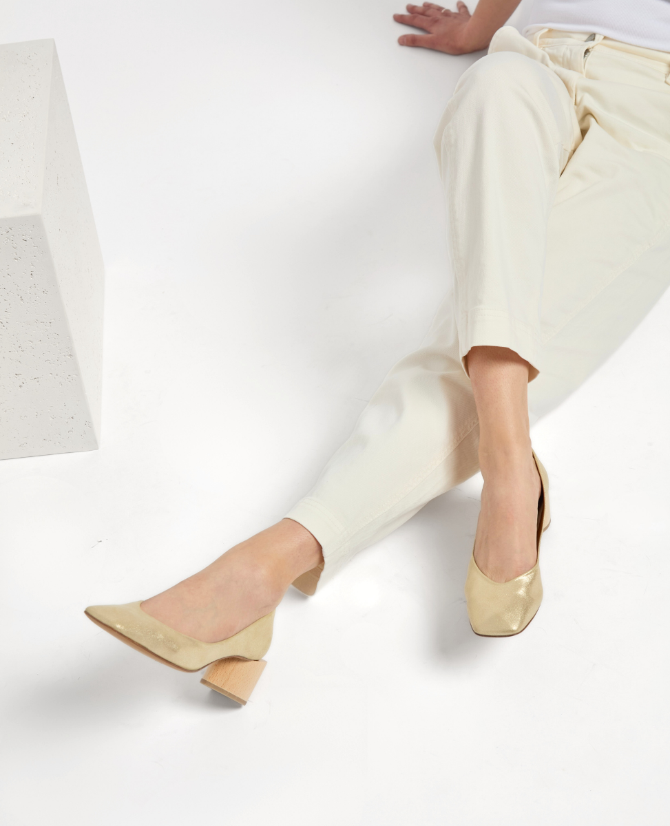 A pair of beige, square-toed pumps with a glossy finish and medium-height wooden block heels, worn by a person in light-colored pants.