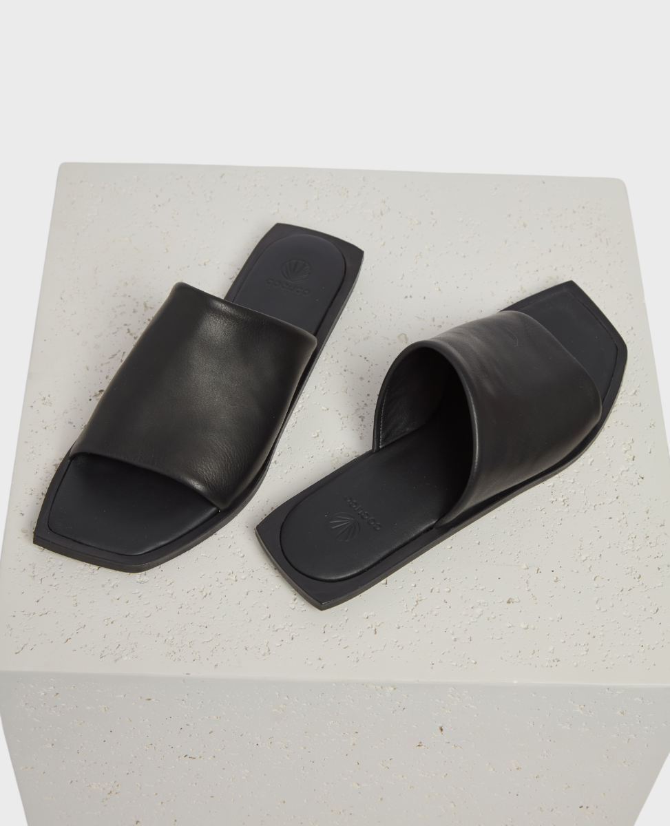 A pair of sleek, black leather slide sandals with a wide strap and square toe design, featuring a smooth texture and minimalist aesthetic.