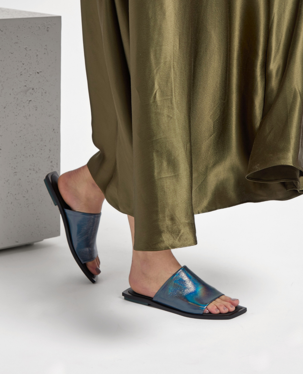 A pair of iridescent blue slide sandals with a sleek, smooth finish, featuring a wide strap across the foot and a flat black sole, worn by a person in a flowing olive-green skirt.