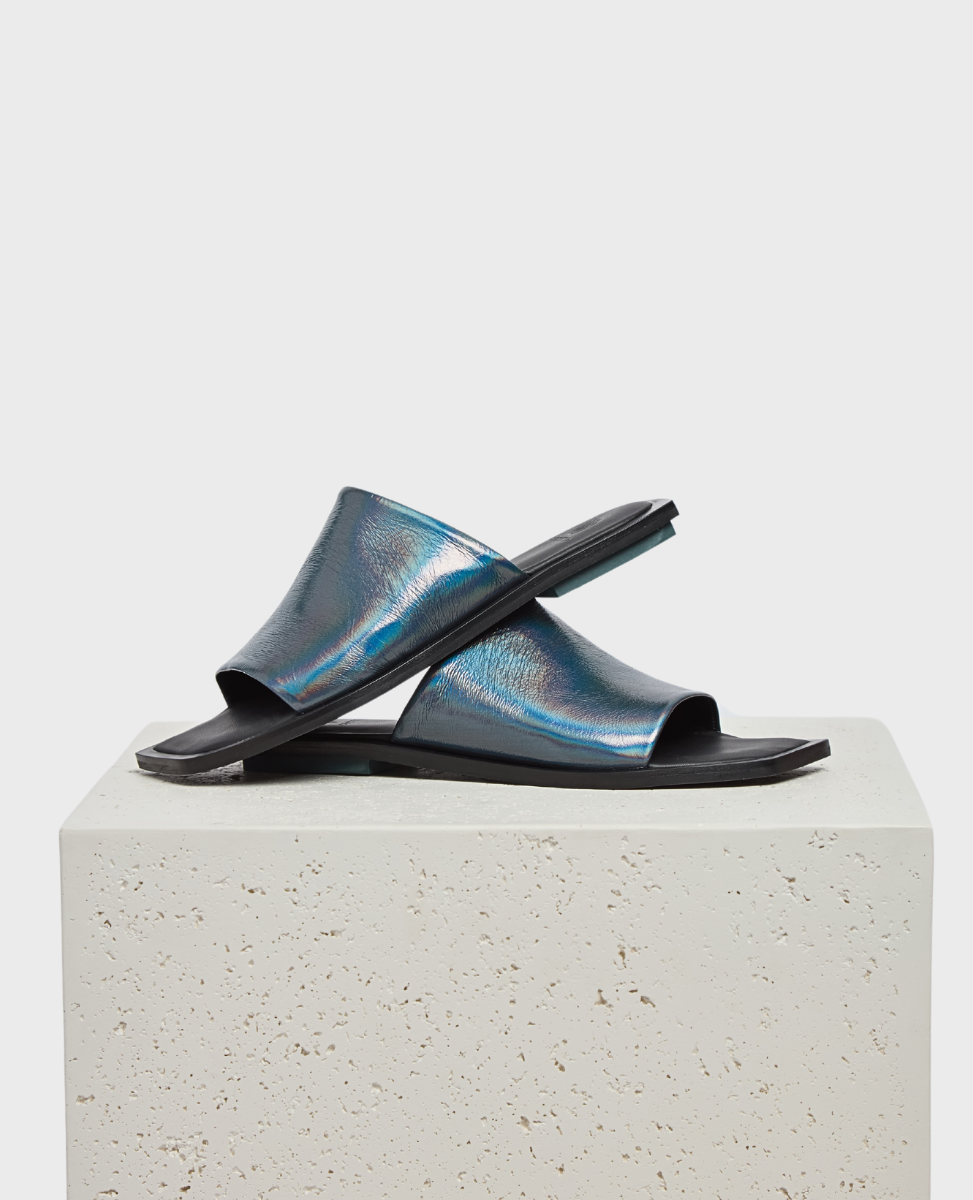 A pair of sleek, metallic blue slide sandals with a glossy finish, featuring a wide strap and flat black sole, displayed on a textured stone block.
