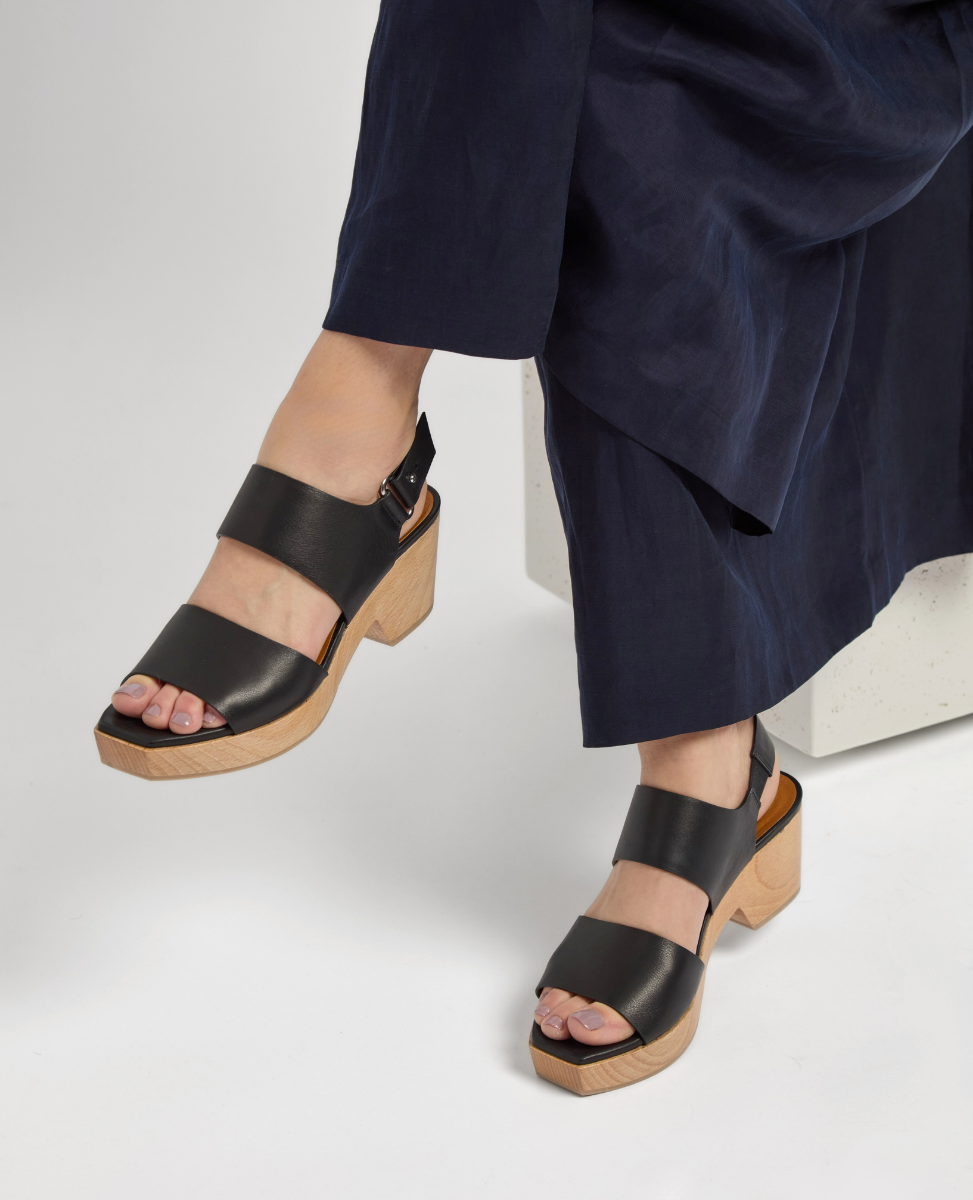 Black leather sandals with a wide strap design, featuring a chunky wooden block heel and an open toe, worn by a person in a dark blue dress.