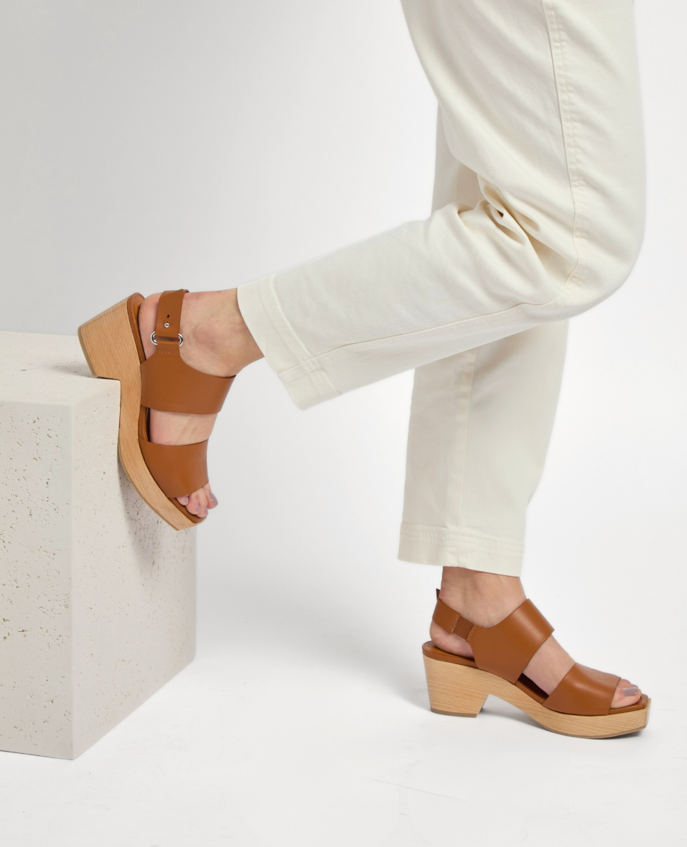 A pair of tan leather sandals with a chunky wooden heel, featuring an open toe design, adjustable ankle strap with a silver buckle, and a smooth finish, worn by a person in light-colored pants.