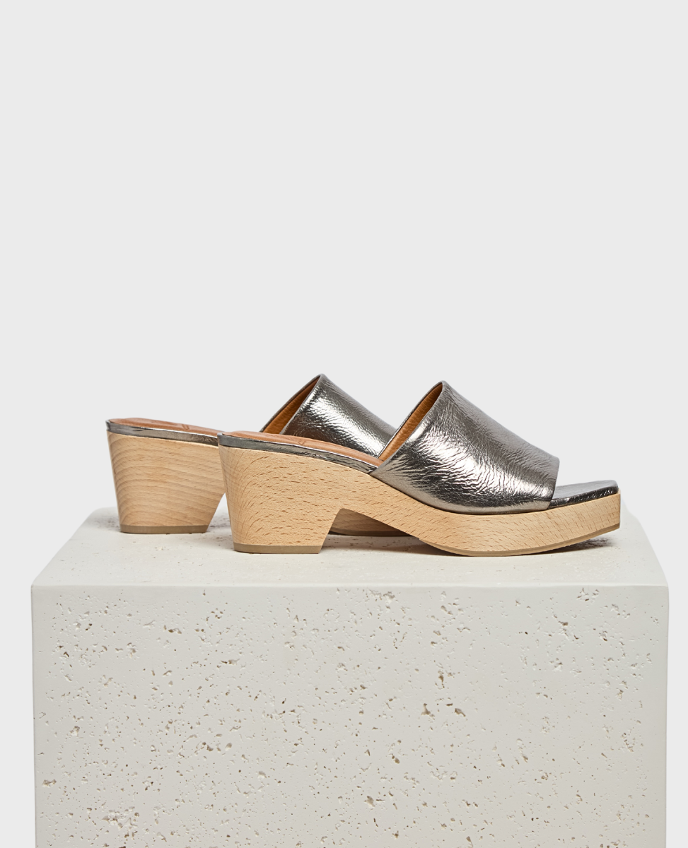 A pair of open-toe mule sandals featuring a metallic silver leather upper with a textured finish, set on a light wooden block heel and platform with a smooth surface.