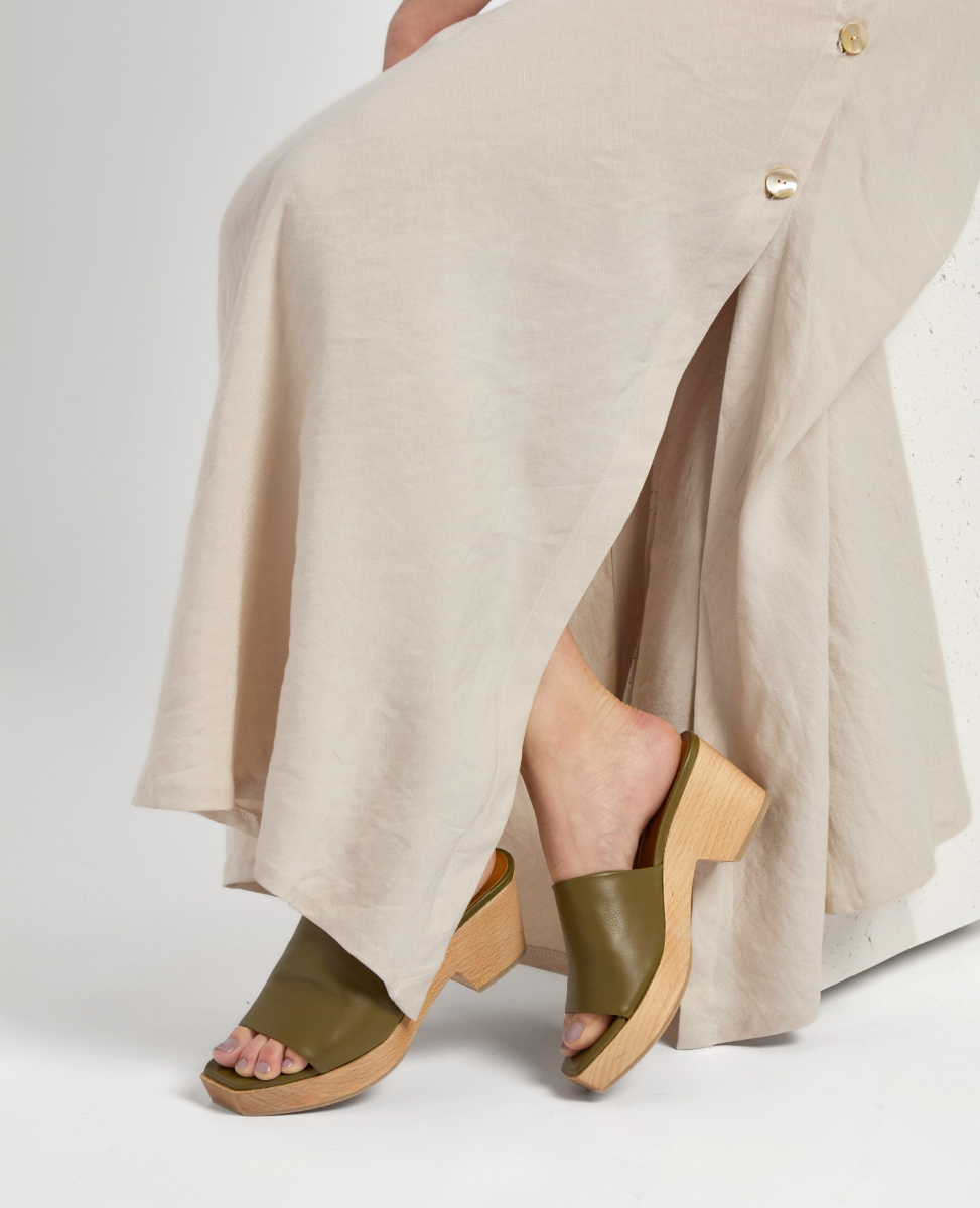 A pair of olive green, open-toe mules with a wide leather strap and a chunky wooden block heel, worn by a person in a flowing beige dress.