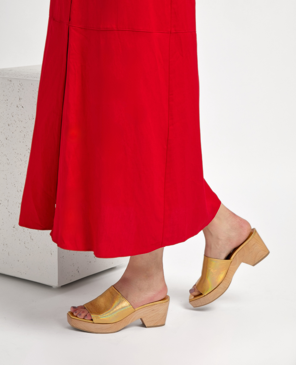 Golden open-toe mules with a shiny finish, featuring a wide strap and a wooden block heel, complementing a vibrant red skirt.