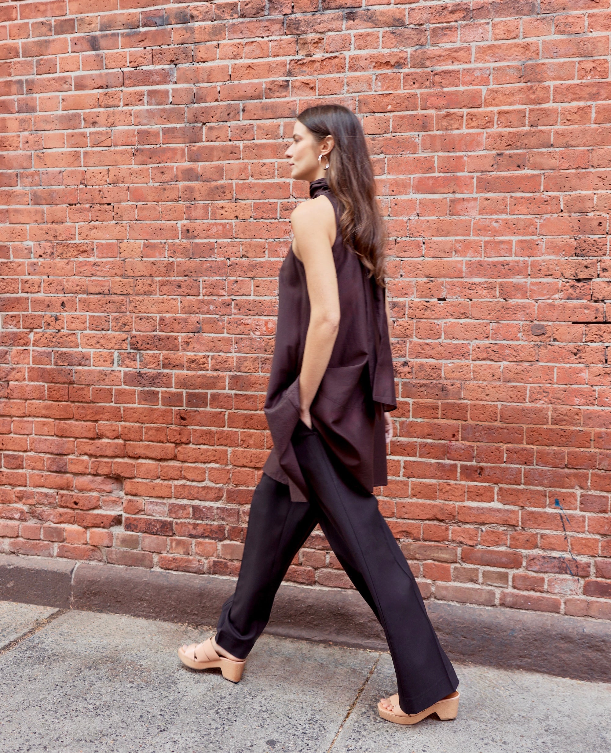 Woman walking against a brick wall wearing a dark sleeveless top and pants.