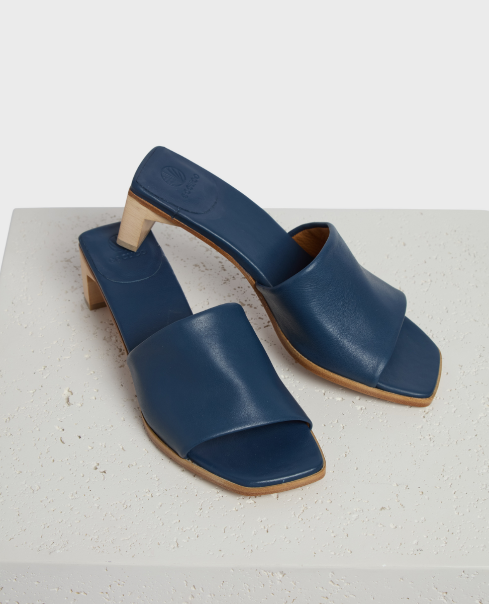 A pair of elegant blue leather slide sandals with a square toe, featuring a low wooden block heel and a smooth, minimalist design.
