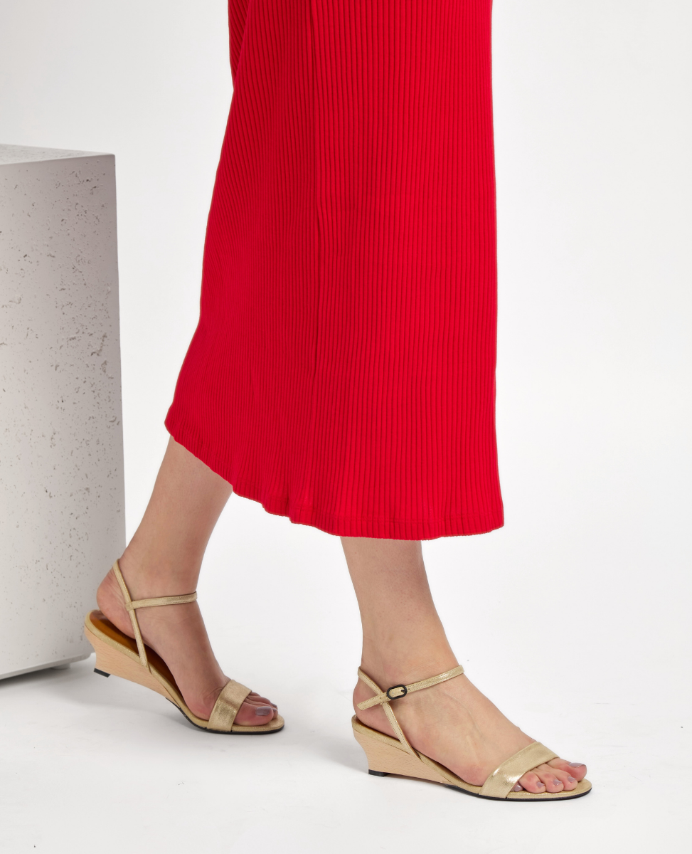 A pair of elegant, gold-colored wedge sandals with thin straps and a small buckle, featuring a smooth texture and a modest heel, complementing a vibrant red, pleated skirt.