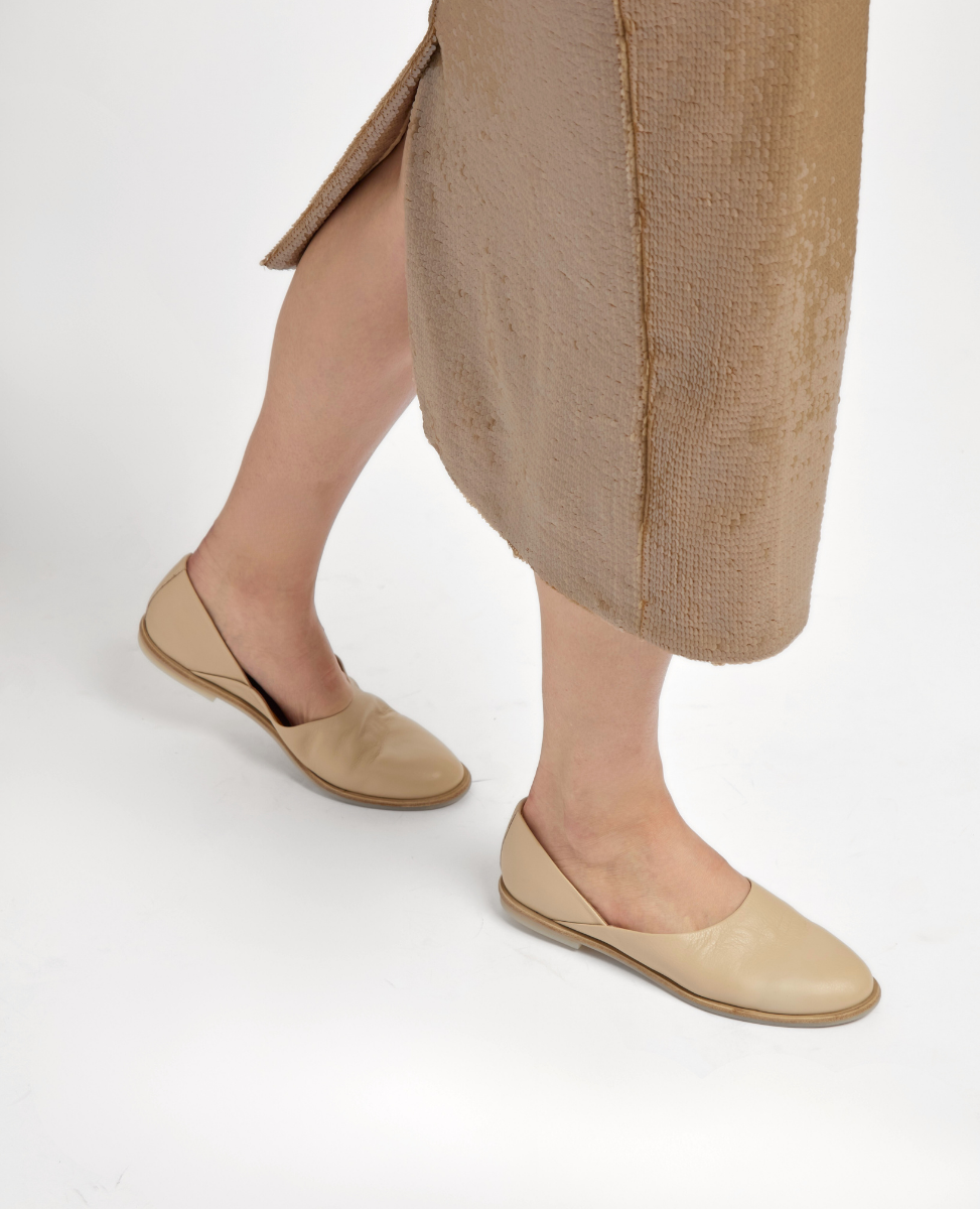 Beige slip-on flats with a smooth, matte finish and a simple, rounded toe design, worn with a textured, light brown skirt.
