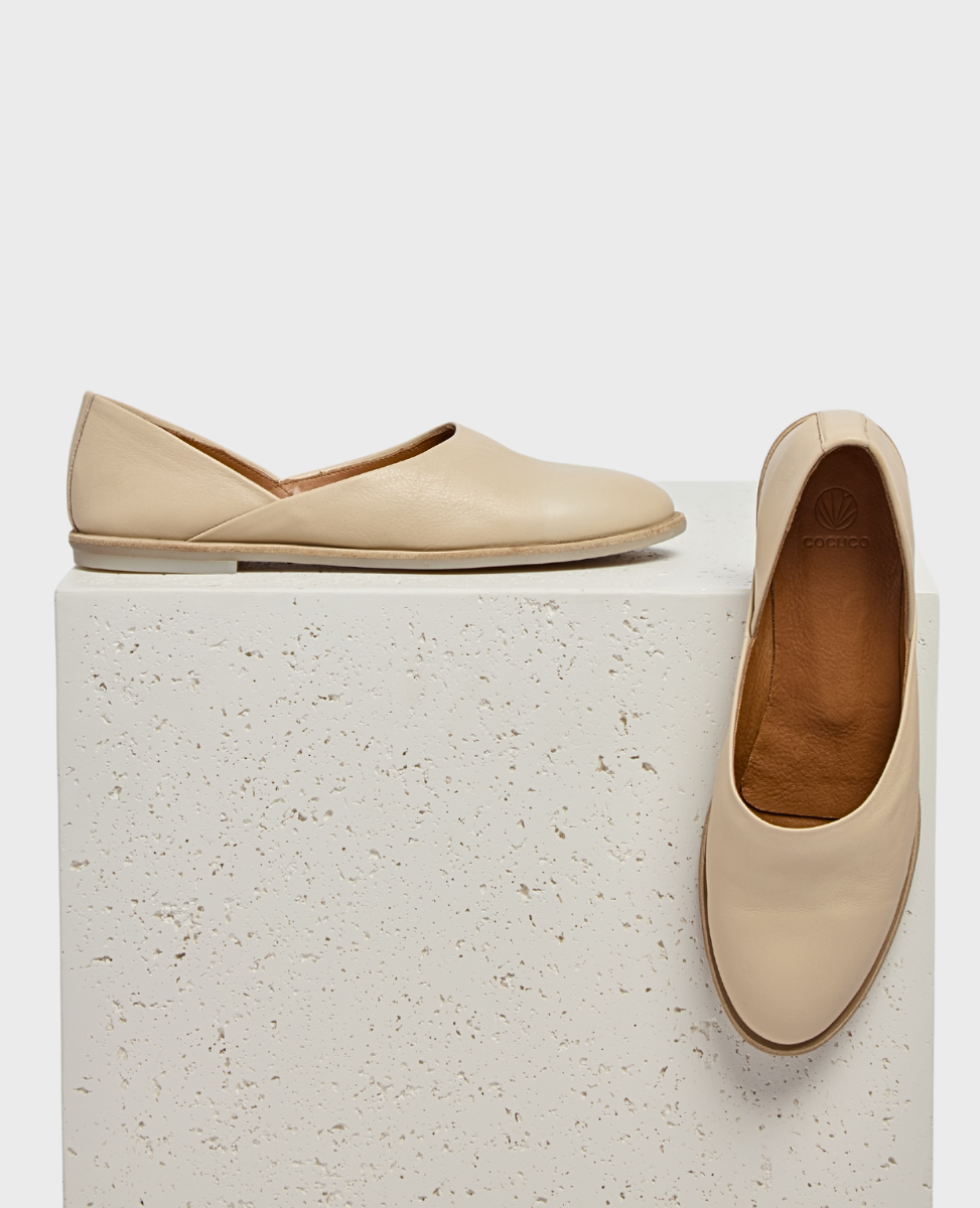 A pair of sleek, beige leather flats with a smooth texture, featuring a minimalist design and a slightly pointed toe, displayed on a textured white surface.
